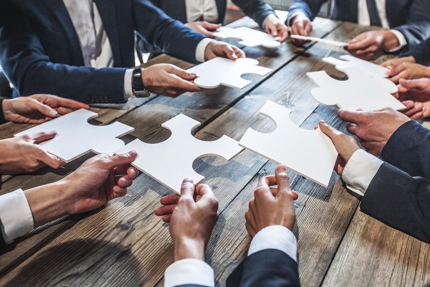 Unternehmer sitzen an einem Meetingtisch und halten Puzzleteile in den Händen